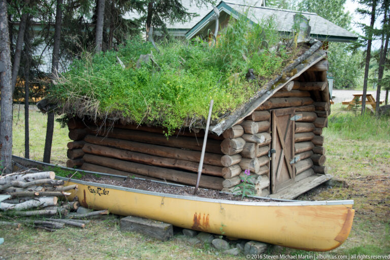 A yellow canoe rests beside a low log shelter with a grass-covered roof, among trees and cut branches.