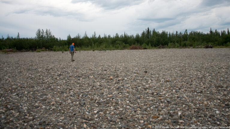 A lone person walks across a broad gravel river bar toward a distant line of shrubs and trees under cloudy skies.