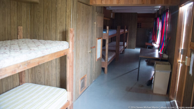 A narrow wood-paneled bunkroom has built-in beds along both sides of a central aisle.