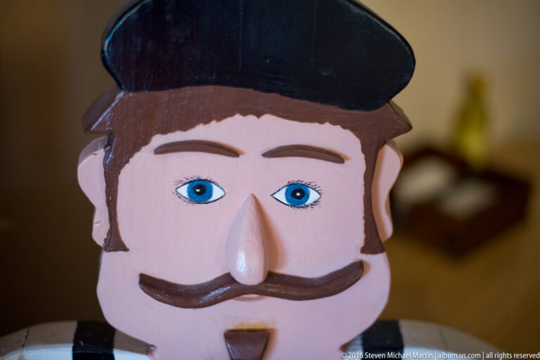 A painted wooden face has blue eyes, a curled dark moustache, and a black cap.