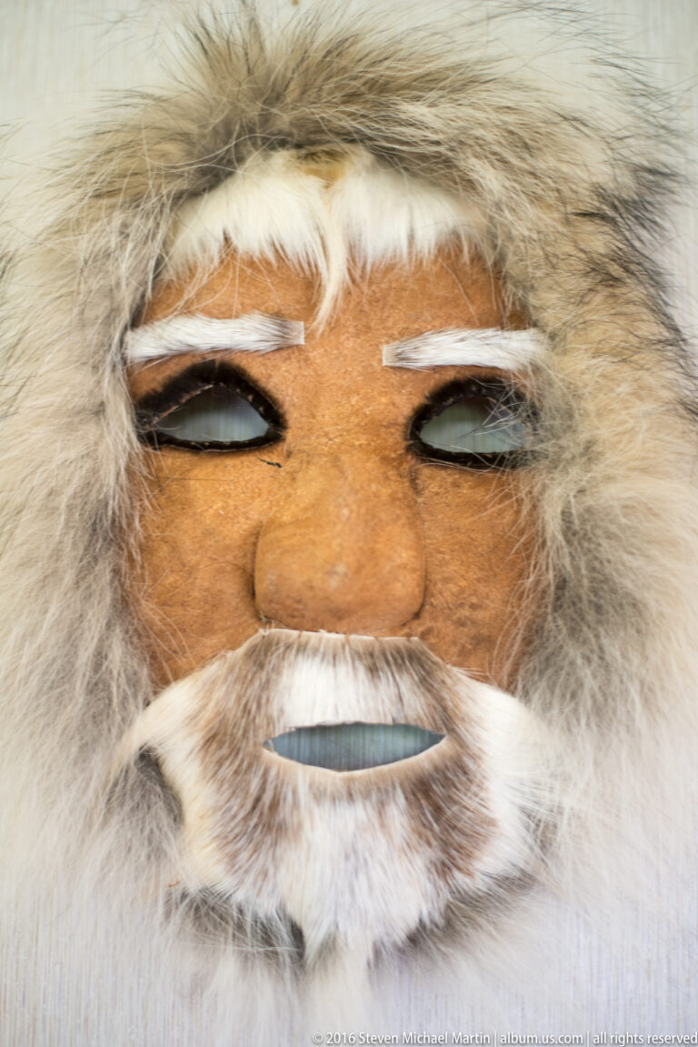 A carved face with dark eye openings, white moustache, and beard is framed by pale fur.
