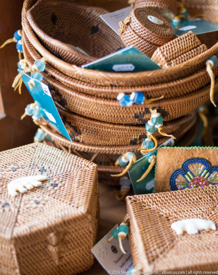 Coiled woven baskets with blue and white decorations are stacked on a tabletop among smaller basketry items and tags.