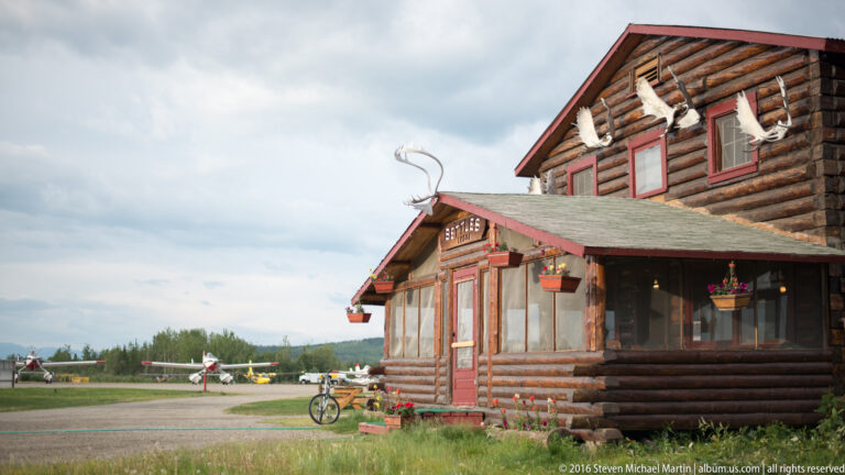 A log building with red trim and antler decorations stands beside an airfield, with small planes and a bicycle nearby.