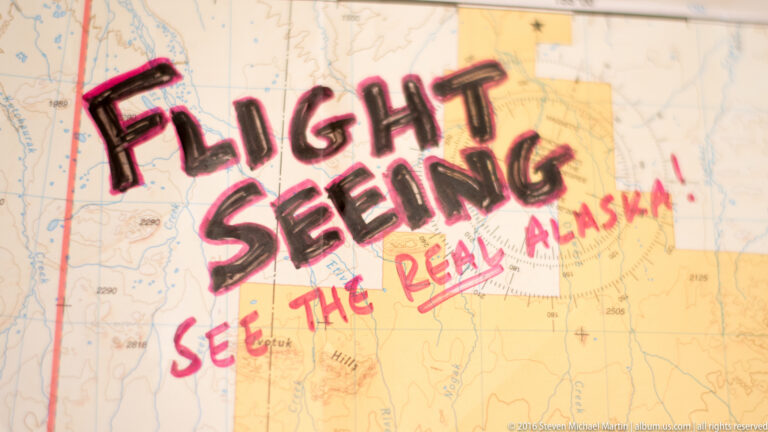 Black and pink handwritten flightseeing slogan crosses a pale map of Alaska, with the lettering filling most of the frame.