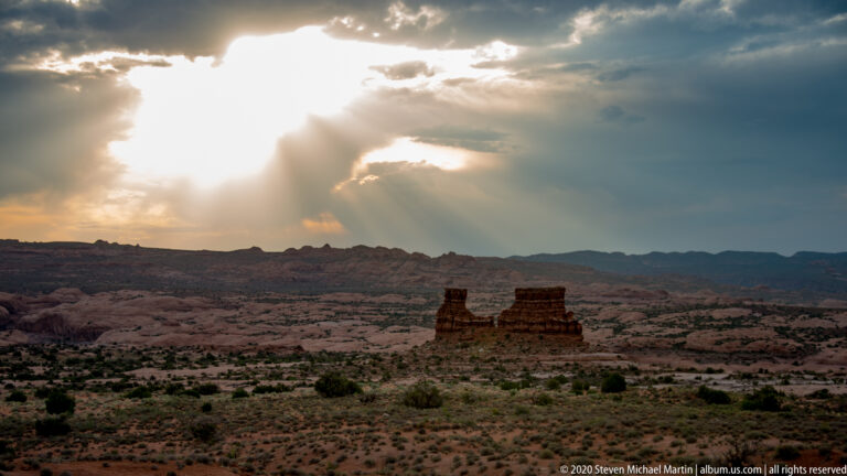 Sunbeams break through clouds above a silhouetted sandstone formation and desert plateau.