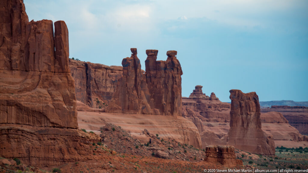 Three narrow sandstone towers stand before layered cliffs and smaller distant buttes.