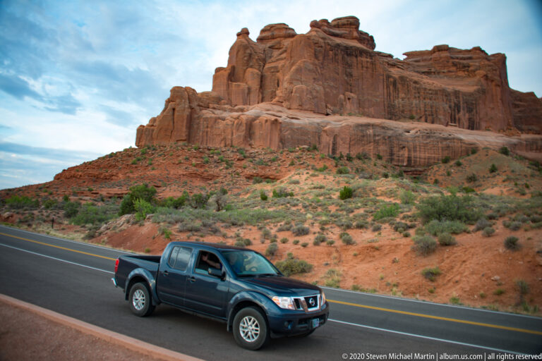 A pickup truck passes below a broad red sandstone cliff dotted with desert shrubs.