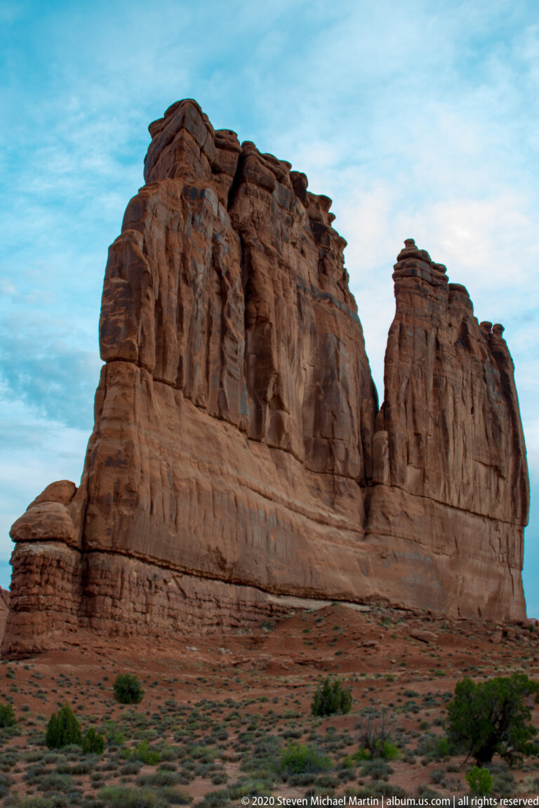 A high red sandstone wall with vertical ribs rises above sparse desert vegetation.