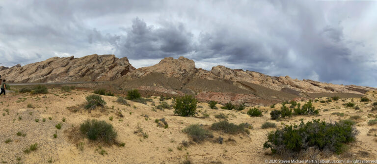 A pale eroded ridge lies beneath storm clouds, with a visitor beside the road at left.