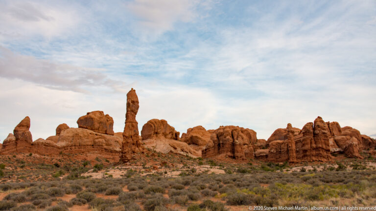 A narrow sandstone pinnacle rises above a long ridge of rounded red rock fins.