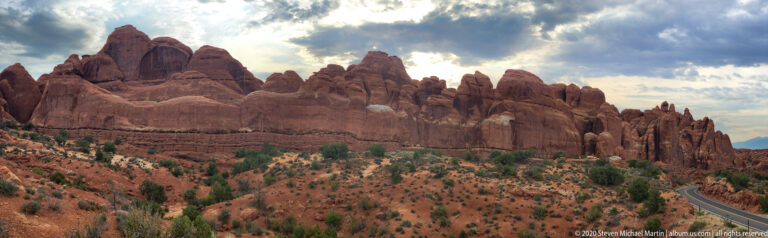 A jagged sandstone ridge rises above a shrub-covered slope, with a road at far right.