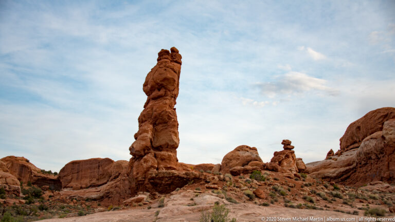 A tall sandstone pillar rises among rounded rock mounds and pale slickrock.