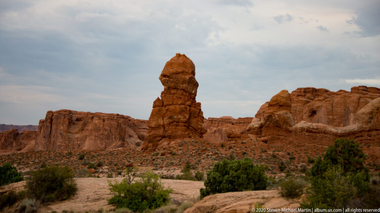 A large sandstone boulder balances on a narrow pedestal before red cliffs.