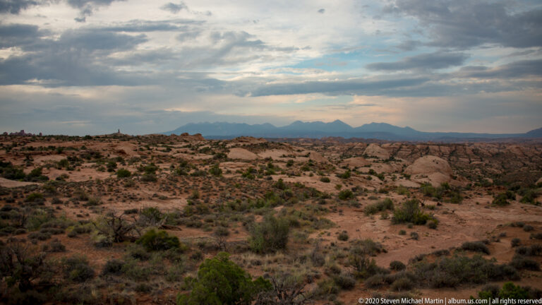 Sparse desert scrub and exposed rock extend toward distant mountains under layered clouds.