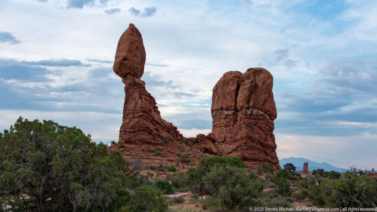 A capstone-topped pillar and a rounded sandstone mass rise above juniper shrubs.