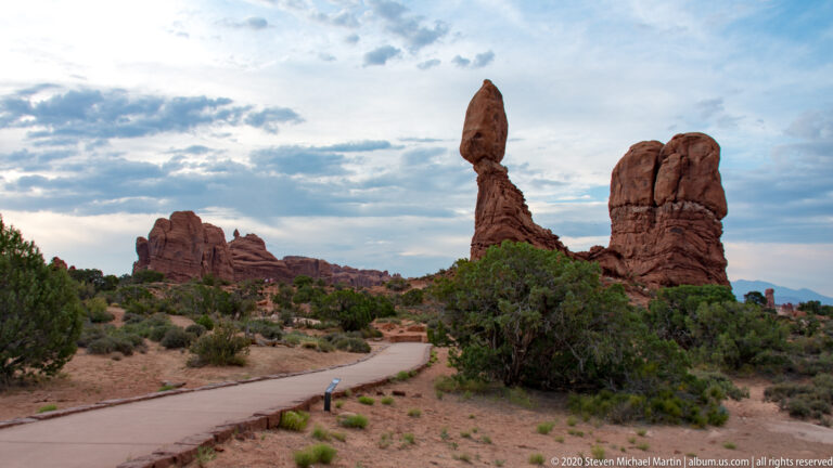A paved path approaches a capstone-topped rock pillar beside a taller sandstone mass.