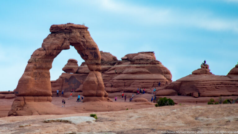 Visitors stand beneath Delicate Arch, a freestanding sandstone span above an open slickrock basin.