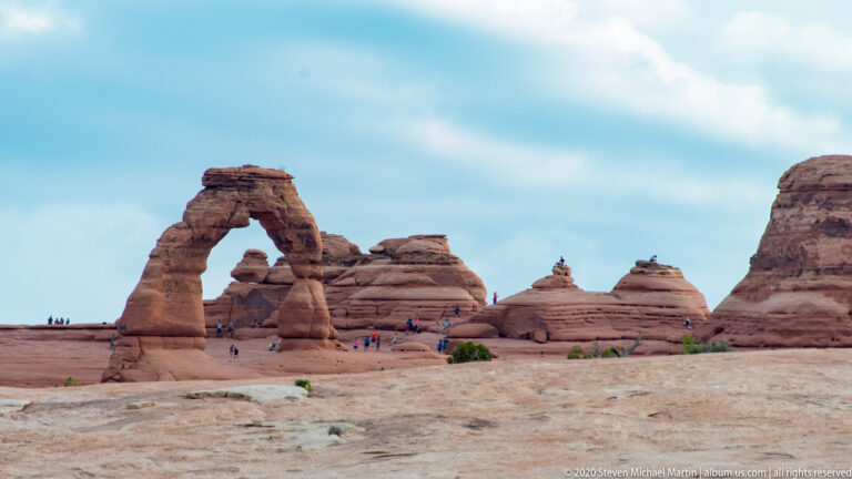 Delicate Arch appears at left above a broad slickrock shelf, with visitors near its base.