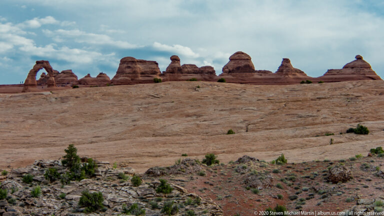 A small arch and rounded sandstone domes line the crest of a broad slickrock slope.