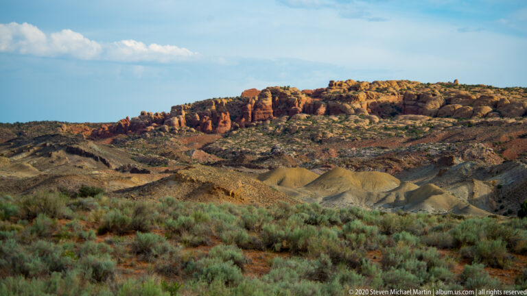 A sunlit red sandstone ridge rises beyond an eroded basin dotted with shrubs.