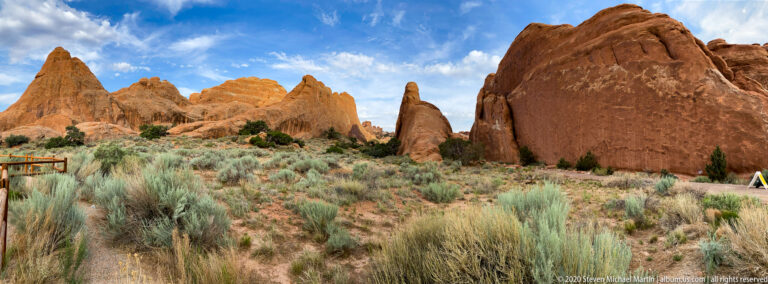 Tall red sandstone formations stand beyond desert shrubs, with a low railing at left.