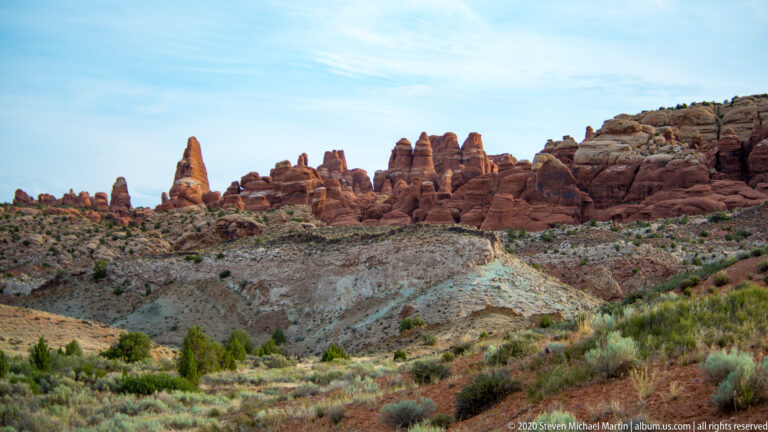 Red sandstone fins cluster along a ridge above pale rock, a shallow wash, and sparse shrubs.