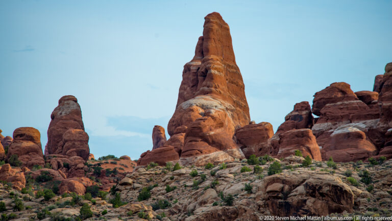 A narrow sandstone fin towers over shrubs and shorter red-rock formations beneath a clear sky.