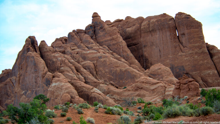 Closely spaced red sandstone fins rise above shrubs and dry grass on a sloping desert floor.