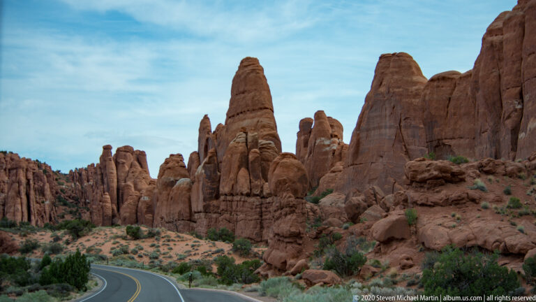 A paved road curves past a tall sandstone fin and a row of red rock towers.