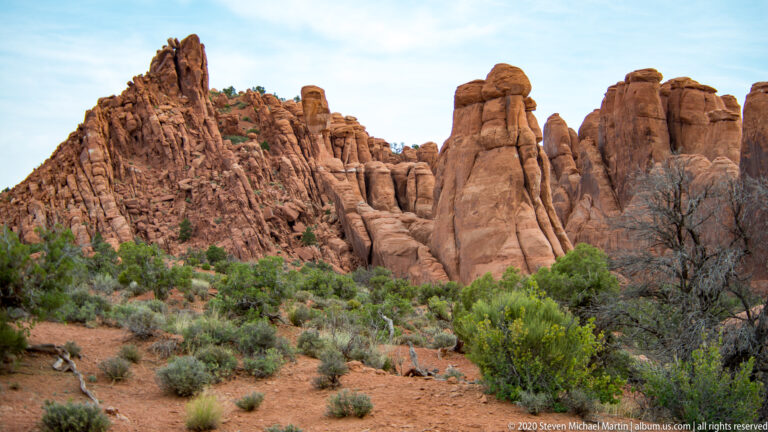A serrated red sandstone ridge rises beyond green shrubs and dry grass.