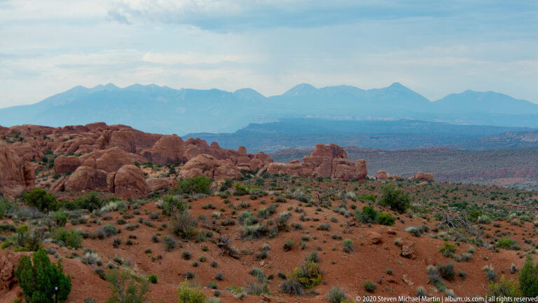 Red sandstone formations and shrubs overlook a hazy valley and distant blue mountain range.