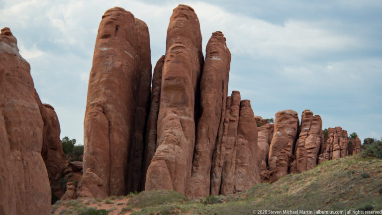 A row of narrow sandstone fins recedes across a scrub-covered slope beneath a cloudy sky.