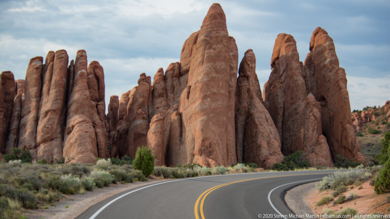 A curving two-lane road passes beneath a tall sandstone tower and a row of red fins.