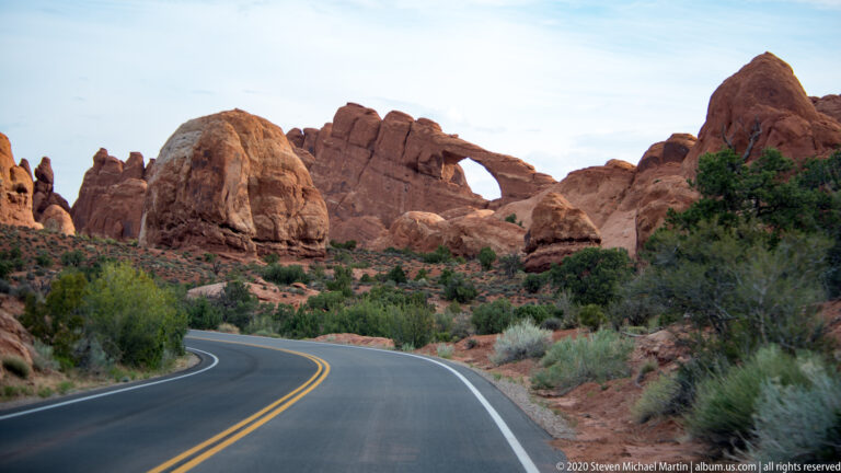 A paved road curves toward a natural arch framed by red sandstone formations and desert shrubs.
