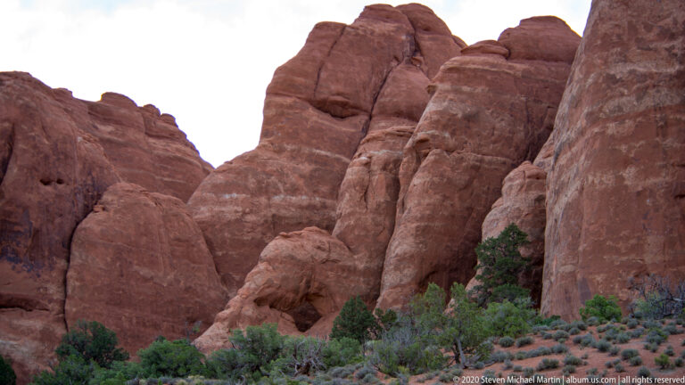 Rounded red sandstone fins overlap above shrubs and reddish soil in a shallow basin.