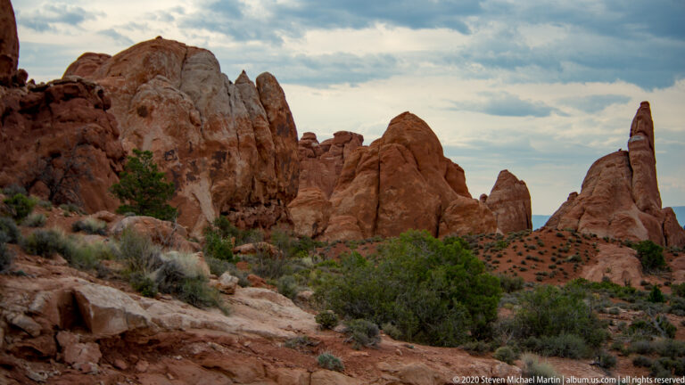 Red fins and rounded sandstone towers extend across a scrub-covered basin beneath clouds.