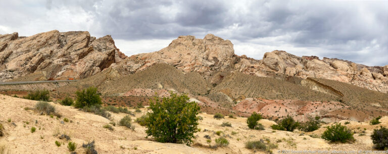 Pale sandstone ridges rise above scrubland, with a road crossing the slope beneath dark clouds.