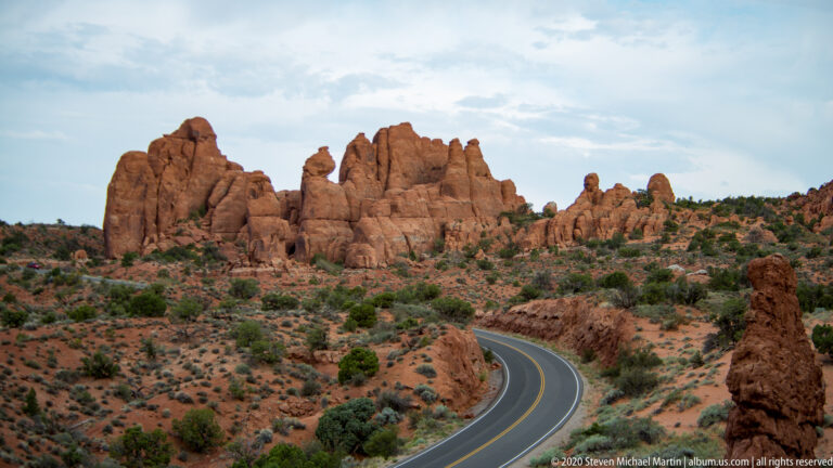 A curved park road passes beneath red sandstone towers and low desert shrubs.