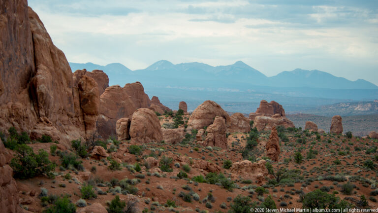 A sandstone wall frames scattered red formations, a valley, and distant blue mountains.