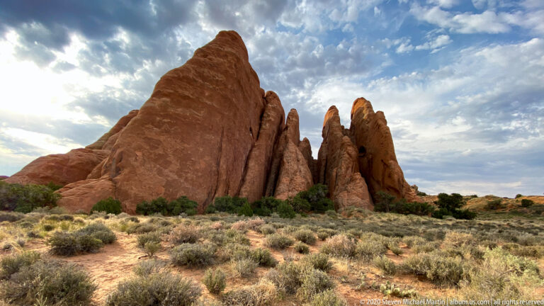 A massive sandstone fin stands above scrub, with narrower spires behind it.
