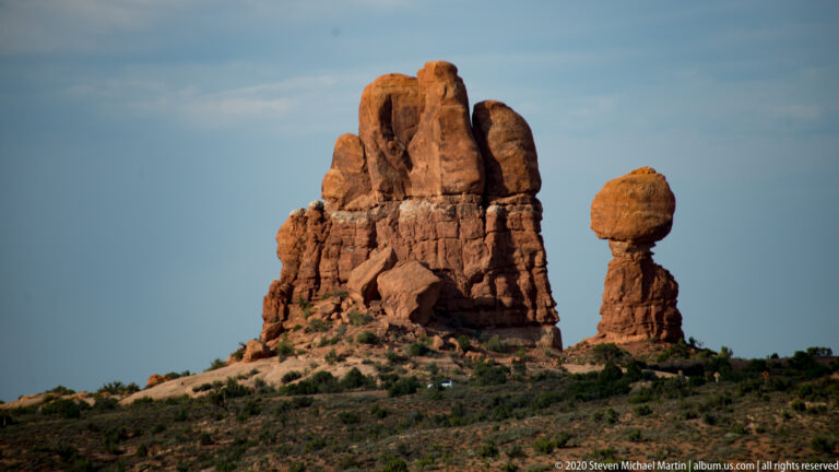 A broad sandstone tower and a smaller pedestal topped by a boulder rise against clear sky.