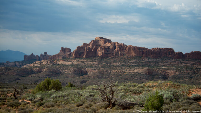 A distant layered sandstone mesa rises beyond sage-colored scrub beneath a hazy sky.