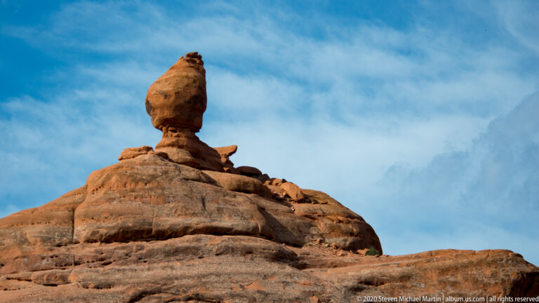 A rounded boulder crowns a narrow rock column on a broad sandstone dome.