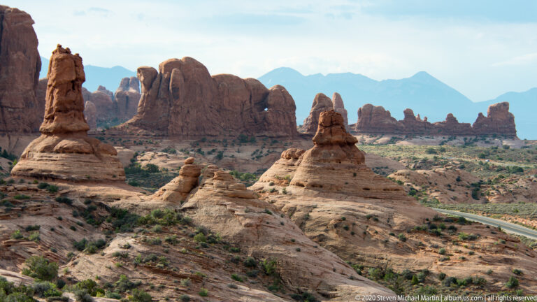 A curved road crosses pale slickrock beneath scattered sandstone formations and distant mountains.