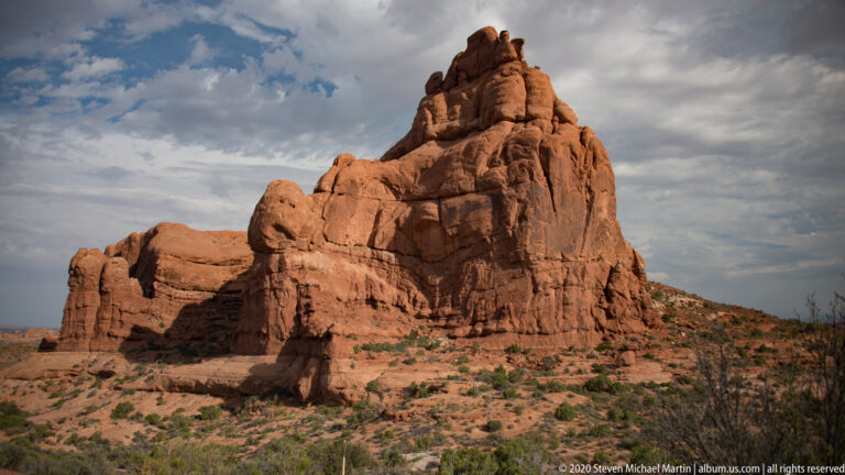 A jagged sandstone tower rises beneath dark clouds, with shrubs along its base.