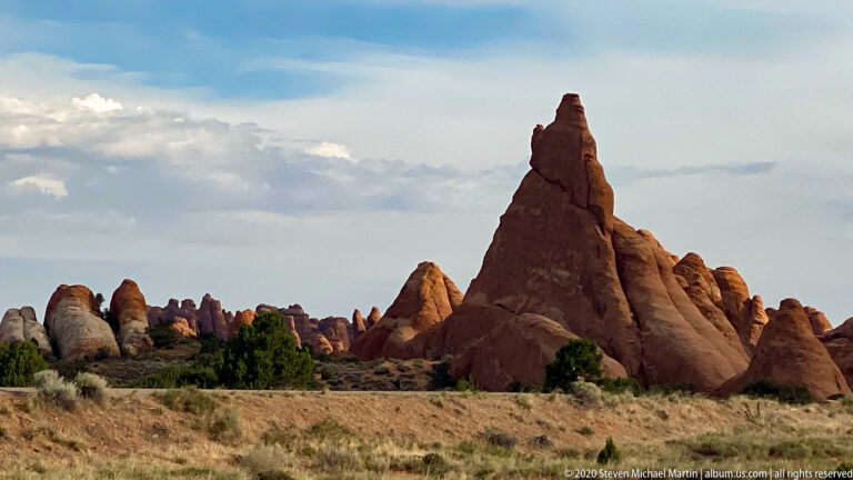 A slender red sandstone pinnacle rises above a low ridge and open grassland.