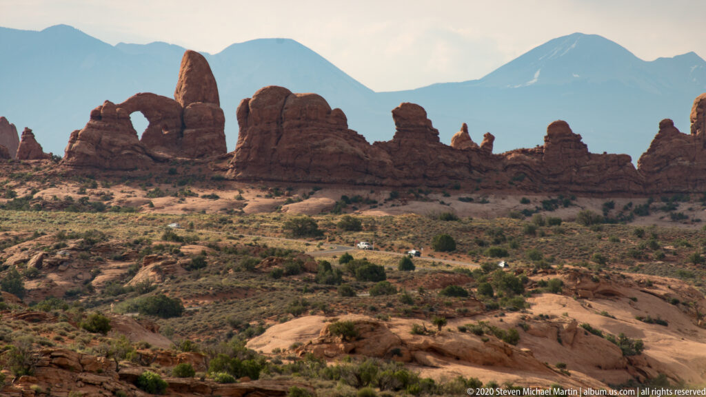 Several arch openings punctuate a sandstone ridge with blue mountains beyond and slickrock in front.