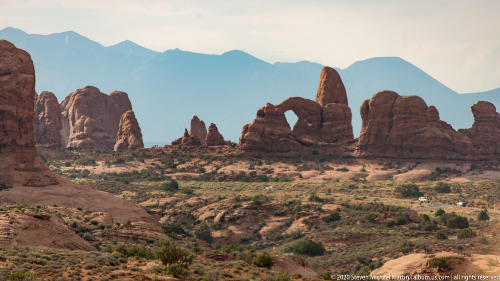Wide desert view of sandstone arches and pinnacles above scrub-covered terrain, with distant mountains behind.