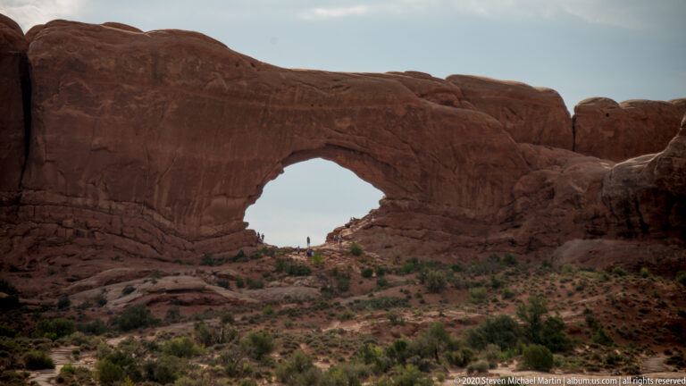 Shadowed sandstone arch with tiny visitors beneath its broad opening and scrub-covered slopes around it.