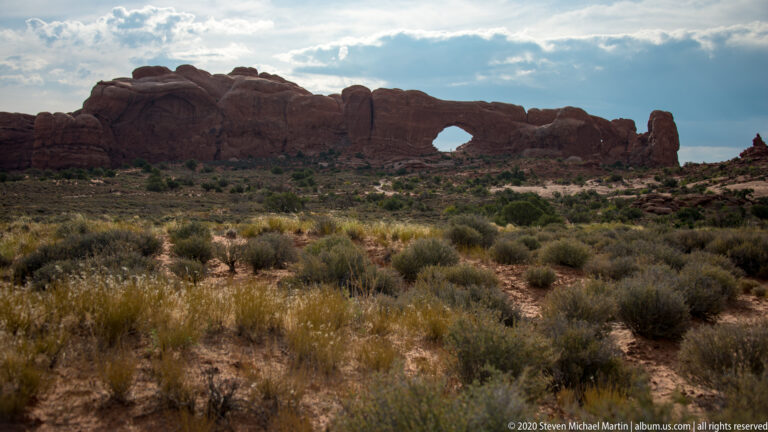 Distant arch opening in a sandstone wall beyond dense sagebrush and grasses.